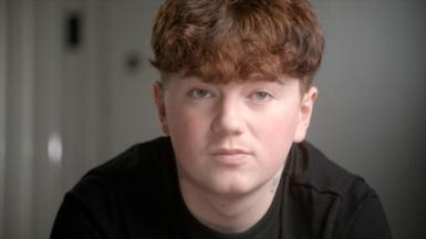Close-up of Alex's face in the foreground, with short curly ginger hair and a dark top, looking towards the camera. A small neck tattoo is visible above the collar. The background is an indoor setting with a plain wall and a blurred door handle or fixture on the left. No bags, bins, or visible markers are present.
