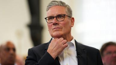 Keir Starmer looking into the distance with a nervous expression. He is inside a room which is unseen, nd wearing a black suit jaclket and white shirt with no tie