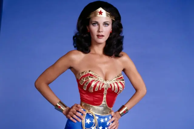 Lynda Carter w swoim charakterystycznym kostiumie Wonder Woman na zdjęciu promocyjnym wykonanym 1 sierpnia 1977 roku. Strój składa się z czerwono-złotej góry oraz niebieskich krótkich spodenek z białymi gwiazdami. Na czole ma złotą trójkątną koronę z czerwoną gwiazdą, na nadgarstkach srebrne mankiety z czerwonymi gwiazdami, a z pasa zwisa złota lina zwana Lassem Prawdy.