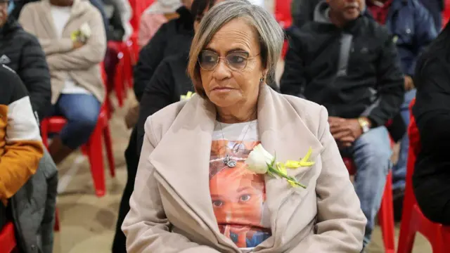 Amanda Smith-Daniels yicaye mu rukiko. Yambaye ikoti rirerire ry'ibara rya 'beige', imbere yambayemo umupira uriho ifoto ya Joshlin Smith. Ku ikola ry'ikoti, hatendetseho ururabo rw'iroza rw'umweru. Yicaye ku ntebe ya plastike y'ibara ry'umutuku, ari kumwe n'abandi bantu bicaye, mu rukiko rukuru rw'i Western Cape, mu gace ka Saldanha Bay, muri Afurika y'Epfo, ku itariki ya 29 Gicurasi (5) mu 2025, bategereje ko isomwa ry'urubanza ritangira. 