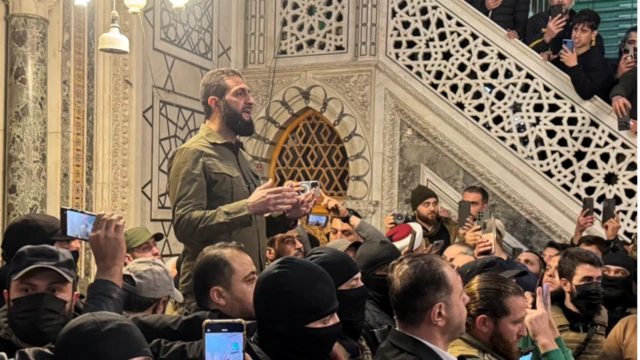 Man in an olive green jacket standing tall above a group of men inside a mosque