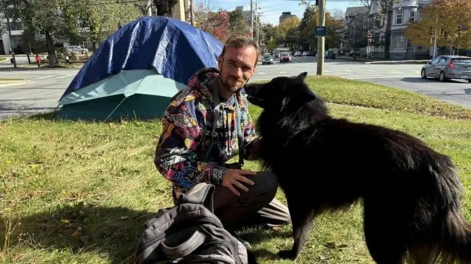 Homem agachado ao lado de cachorro, em gramado com barraca ao fundo, em zona urbana