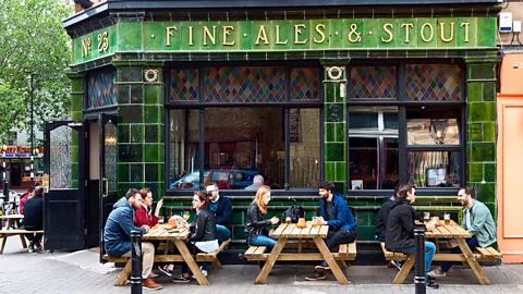 People sit outside a green-tiled pub in London on wooden benches with gold lettering reading 'Fine Ales and Stout' (Credit: Alamy)