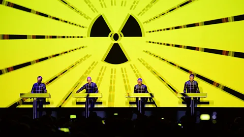 The four members of Kraftwerk performing on stage with a radioactive symbol against a bright neon yellow screen behind them (Credit: Getty Images)