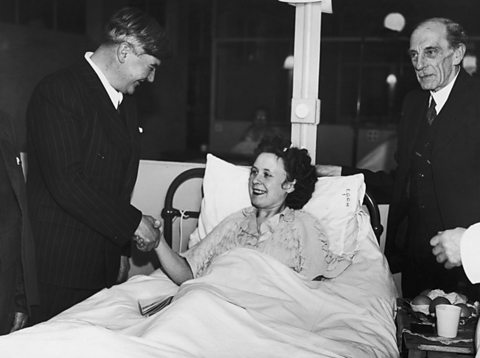 Aneurin Bevan smiles and shakes the hand of a female patient in a hospital bed.