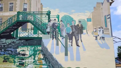 Tim Carroll A newly repainted mural with a prominent bridge on the left and a number of figures on the right. Ken White can be seen under a flat cap with his arms crossed.